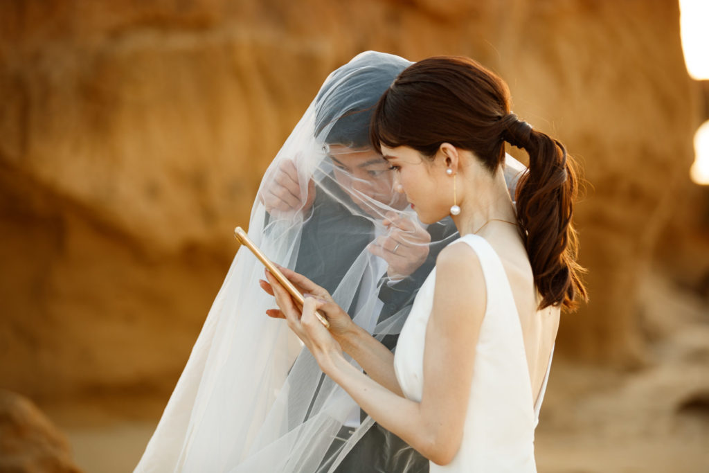 奈多海岸の第三崖の前でタブレットを囲みながら、撮影したばかりの写真を笑顔で確認する新郎新婦の様子