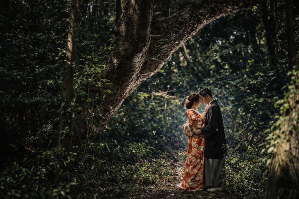 奈多海岸の深い森の中で、新郎新婦が和装姿で寄り添う前撮り写真。差し込む光に包まれ、幻想的な雰囲気の中で向かい合う二人の姿を捉えた一枚。